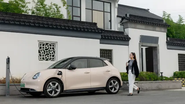 Cao Mengyi holds a coffee cup while standing in front of her car Ora Good Cat in Suzhou. She looks into the camera. A tree-filled park and skyscrapers are visible in the background