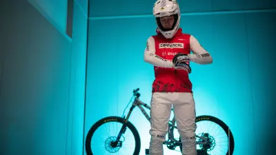 Mountain biker in red and white gear standing in front of a bike in a blue-lit room.