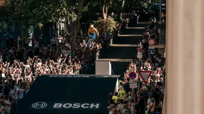 Mountain biker airborne above a ramp on a city street surrounded by spectators.