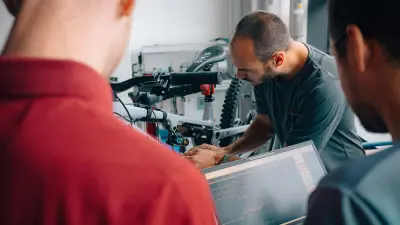 Person working on a mountain bike while two people observe, with a laptop in the foreground.