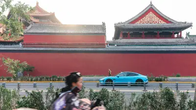 A blue car drives past a red wall with traditional Chinese architecture.