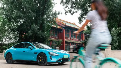 A sleek blue electric car parked in front of a traditional Chinese building. A blurred figure rides a bicycle past the car.