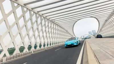 A blue sports car drives on a modern bridge with a white lattice structure.