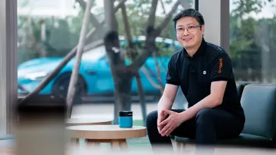 Zhenyu Huang sitting on a chair with the Xiaomi SU7 in the background.