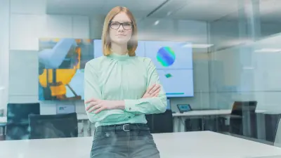 Sarah Fischbach leans on her desk with her arms folded. In the background is a large screen with diagrams.