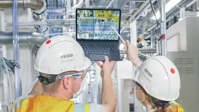 Two associates wearing helmets and protective clothing peer at a monitor with numerous cables and circuits.