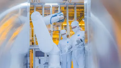Two associates standing in the fab wearing white coveralls gaze at a white robot arm.