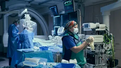 Jessica Rama in blauer Krankenhausbekleidung mit Kopftuch und Gesichtsmaske vor einem Krankenbett und Medizintechnikgeräten