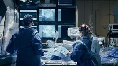 Zwei Menschen in blauer Krankenhausbekleidung vor einem Krankenbett und vielen Monitoren