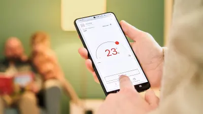 Melanie Wingerter blickt auf ein Smartphone, auf dem die App der Wärmepumpe aufgerufen ist. Im Hintergrund spielt David Wingerter mit den Kindern.