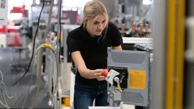 A young woman assembles a protective plate on the back of a heat pump in a factory building.