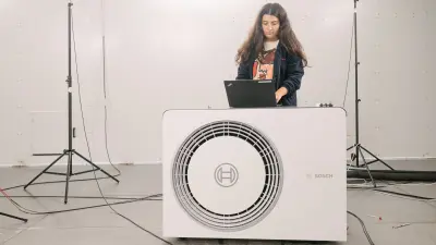 Eine Bosch-Mitarbeiterin mit Laptop steht im Schalllabor des portugiesischen Standorts Aveiro hinter einer weißen Wärmepumpe.