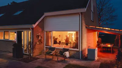 Rechts neben einem Haus in Hasselt in den Niederlanden steht eine Wärmepumpe von Bosch. Es ist Abend und im beleuchteten Wohnzimmer unterhalten sich zwei Menschen.