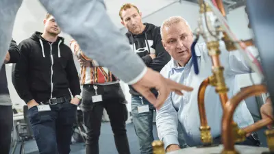 Der Bosch-Trainer für Heiztechnik Gert Brommer kniet bei einer Schulung hinter einer Inneneinheit einer Wärmepumpe und erklärt den zusehenden Teilnehmern die Technik.