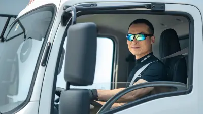 The driver wearing a black t-shirt and sunglasses behind the wheel of the truck shot from the left, smiling at the photographer through the open driver-side window