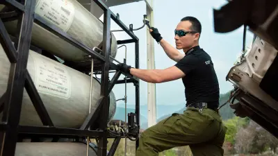 A man with an athletic build wearing a black t-shirt, sunglasses, and gloves looks at the tank below the truck’s tilted cabin