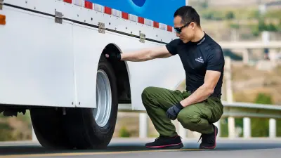 A man with an athletic build wearing a black t-shirt, sunglasses, and gloves squats at the truck’s rear left twin wheel 