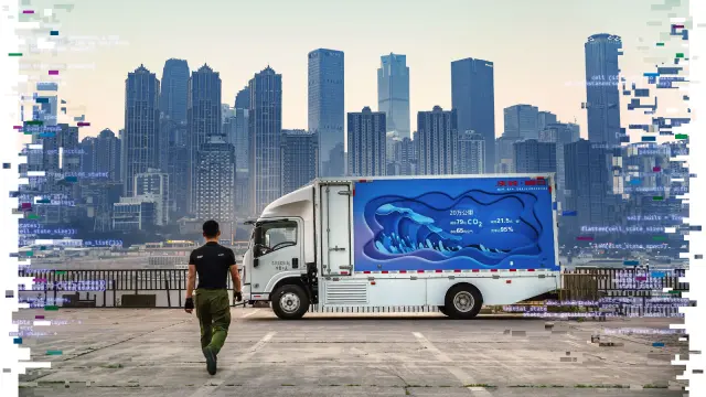 A young man in a black t-shirt, seen from behind, walks toward a blue and white truck parked sideways. On the horizon is the skyline of a metropolis.