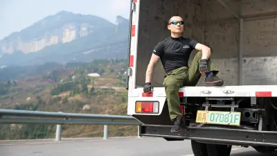 The truck driver Hu Gang, a man with an athletic build wearing a black t-shirt, sunglasses, and gloves, is sitting on the loading area of his truck. A mountain can be seen in the background.