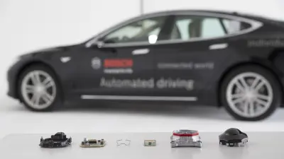 Mehrere Bosch Sensoren und Kameras sind im Vordergrund zu sehen und im Hintergrund ein Testfahrzeug für automatisiertes Fahren.