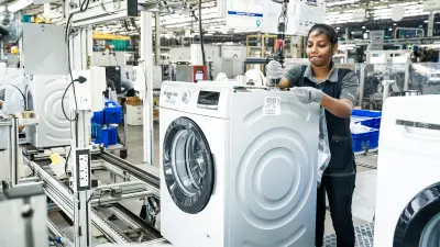 A female factory worker is assembling the top of a washing machine using an electric screwdriver. She is wearing grey gloves and an apron.