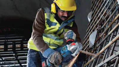 A construction worker on a building site, wearing a yellow helmet, safety goggles, yellow headphones, and a high-vis vest. He has a black beard, is wearing blue and white work gloves, and is operating a blue Bosch angle grinder, which is sending sparks flying.