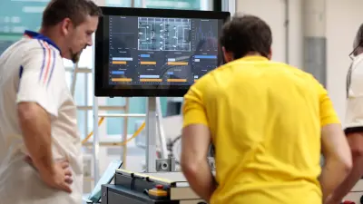 Two men are playing table soccer while in the background one can see a monitor.