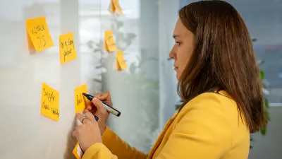 Isabel steht an einem Whiteboard und fügt Haftnotizen hinzu.