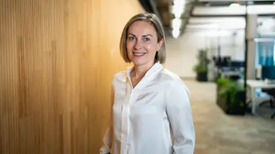 Karolina wears a white blouse, stands in an offi ce and smiles at the camera.