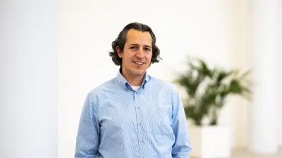 Ozan Demir is standing next to a column. He is wearing a blue and white striped shirt.