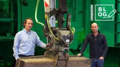 Ozan Demir and Adrian Trachte are standing underneath the arm of the Bosch excavator. Ozan is on the left, Adrian is on the right. Both are touching the excavator while smiling into the camera.