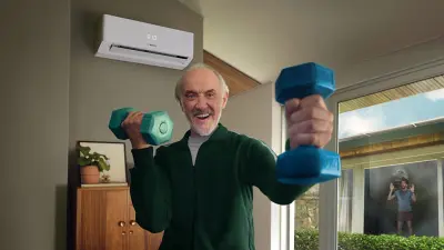 An elderly man is working out with dumbbells in his living room. On the wall behind him is a Bosch air conditioning unit that keeps the temperature comfortable.