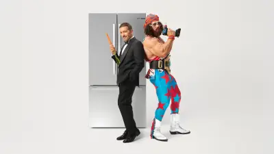Antonio Banderas im schwarzen Anzug und Macho Man Randy Savage im farbenfrohen Wrestling-Kostüm, mit den Rücken aneinander gelehnt, in dynamischer Pose vor einem Bosch-Kühlschrank.