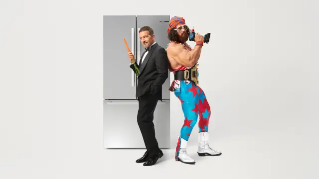 Antonio Banderas im schwarzen Anzug und Macho Man Randy Savage im farbenfrohen Wrestling-Kostüm, mit den Rücken aneinander gelehnt, in dynamischer Pose vor einem Bosch-Kühlschrank.