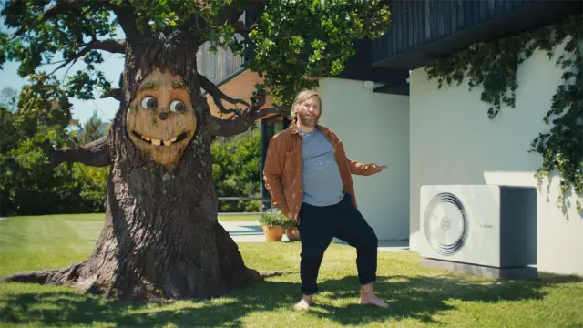 Ein Mann steht neben einem Baum und zeigt auf die Bosch Wärmepumpe neben dem Haus. Der Baum hat ein Gesicht und lächelt dem Mann zu.