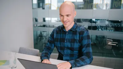 Benjamin wears a chequered shirt and works on his laptop in the office.