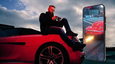 Florian Roschu, world champion in the smartphone racing game "Asphalt 9", sits on the road and leans against a red sports car while playing on his smartphone. Next to him is a large smartphone showing a futuristic super sports car from a game.
