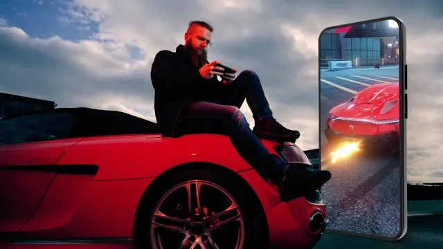 Florian Roschu, world champion in the smartphone racing game "Asphalt 9", sits on the road and leans against a red sports car while playing on his smartphone. Next to him is a large smartphone showing a futuristic super sports car from a game.