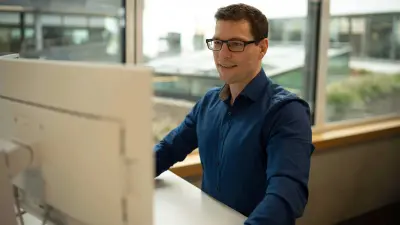 Michael Huis steht an einem Schreibtisch in einer Büroumgebung, er trägt ein blaues Hemd. Vor ihm steht ein Bildschirm, er lächelt, sein Blick ist auf den Bildschirm gerichtet.