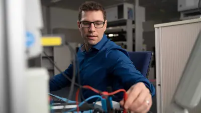 Michael Huis trägt er blaues Hemd, er sitzt auf einem Bürostuhl in einer Laborumgebung, sein Blick ist einem Gerät mit blauen und roten Kabeln zugewandt, er berührt das rote Kabel.
