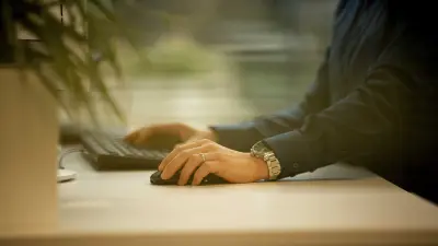 Michael Huis Arme und Hände sind fokussiert. Sie stützen auf einem Schreibtisch, mit der linken Hand hält er eine Computer-Maus, die rechte Hand berührt eine vor ihm liegende, schwarze Tastatur.