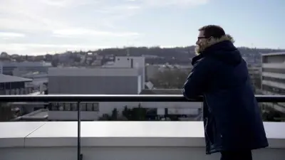 Michael Huis befindet sich in einer Außenumgebung und trägt eine Jacke. Er lehnt seine Arme auf ein Geländer und schaut zur Seite. Im Hintergrund befinden sich der Himmel und mehrere graue Bürogebäude.