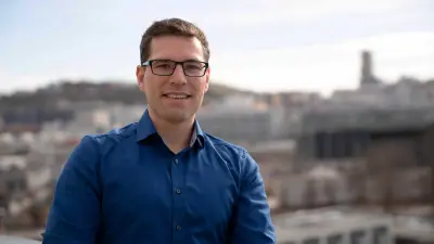 Michael Huis befindet sich in einer Außenumgebung, im Hintergrund sind graue Bürogebäude. Er blickt in die Kamera und lächelt.