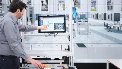 Bosch Research expert Matthias Kayser (left) in front of a monitor at ARENA2036 in Stuttgart and pointing at a screen which shows whether or not the component on the table below – a battery – has been installed correctly. The installation is part of a battery assembly pilot project carried out by Bosch Research together with Mercedes-Benz. The DeepInspect system shows the worker in real time precisely whether or not the component has been installed correctly.