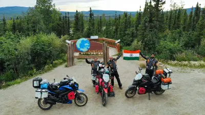 Three men in motorcycle clothing stand with their motorcycles at Arctic Circle and cheer and hold up the Indian flag.