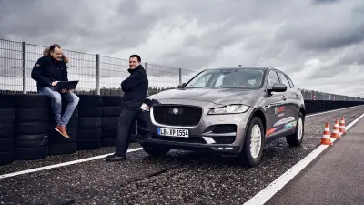 Matthias Birk and Andreas Schulz with a tablet next to a vehicle with an self-driving car camera standing on a test track.