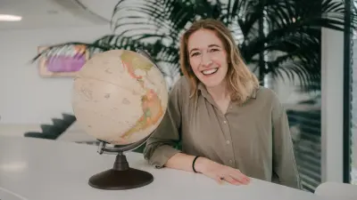 Katharina is standing at a table, leaning on it with her right arm. She is smiling at the camera. On the table next to her is a globe. Behind her is a big green plant.