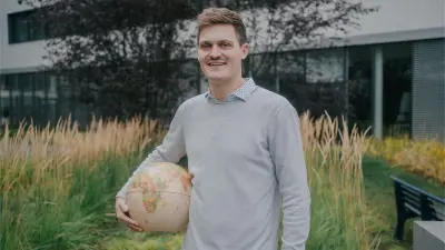 Justus is in an outside area, in the background one can see a building and plants. In his right arm he is holding a globe. He is smiling at the camera.