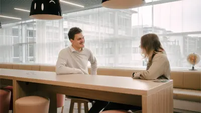 Justus and Katharina are sitting at a long table and are smiling at each other. In the background, one can see an office environment with a long bench, chairs and lamps.