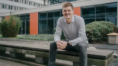 Justus is sitting on a brown wooden bench in an outside area. His arms are crossed as he is smiling at the camera. There is a building and plants in the background.